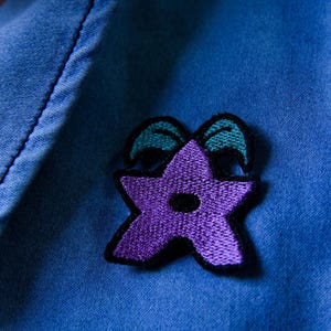 Puede incluir: Parche bordado en forma de estrella púrpura con ribete negro y detalles turquesa, cosido sobre una camisa vaquera azul. El parche es un diseño floral estilizado.
