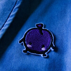Puede incluir: Primer plano de una chaqueta vaquera azul con un parche bordado morado. El parche presenta un dise&ntilde;o de dibujos animados de una criatura redonda y morada con contornos negros y peque&ntilde;os acentos blancos.