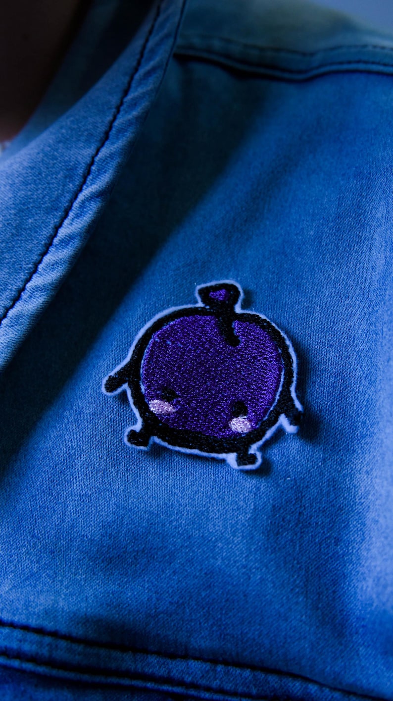 Puede incluir: Primer plano de una chaqueta vaquera azul con un parche bordado morado. El parche presenta un dise&ntilde;o de dibujos animados de una criatura redonda y morada con contornos negros y peque&ntilde;os acentos blancos.