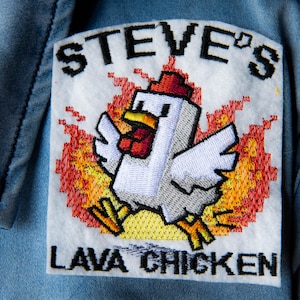 Op de afbeelding: Geborduurde patch met een gepixeld kippetje in vlammen. De patch leest "STEVE'S LAVA CHICKEN" in zwarte tekst. Het is genaaid op een lichtblauwe spijkerjas.