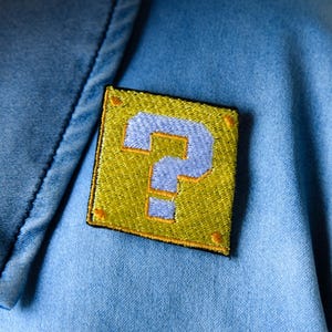 May include: Embroidered patch featuring a yellow question mark block with a blue question mark. The patch is sewn onto a light blue denim fabric. The patch has a black border and orange corner accents.