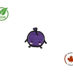 Puede incluir: Parche de fruta morada bordada con una cara linda y contornos negros. El parche tiene un tallo y peque&ntilde;os brazos y piernas. La palabra "VEGAN" est&aacute; en un c&iacute;rculo verde. Hecho en Canad&aacute;.
