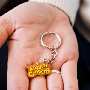 Peut inclure: Un porte-clés brun avec un logo "Animal Crossing" jaune. Le porte-clés est tenu dans la main d'une personne.