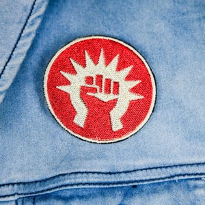 May include: A round, red embroidered patch with a white fist design, surrounded by white starbursts. The patch is affixed to a light blue denim jacket, with visible stitching and a button.