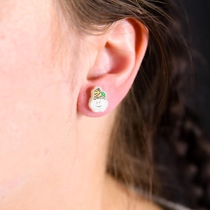 May include: A white pear-shaped earring with a green leaf and two yellow bees on top. The earring is worn in the left earlobe.