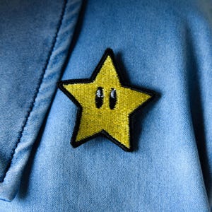 May include: A yellow star-shaped embroidered patch with a black outline and two black eyes, attached to light blue denim fabric. The patch is a popular video game character. The denim fabric has visible stitching.
