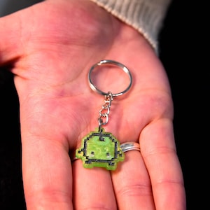May include: A green pixel art alien keyring with a black border and a silver ring. The alien has a simple design with two small eyes and a small mouth. The keyring is held in a hand.
