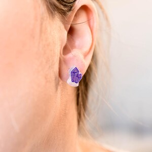 Puede incluir: Primer plano de una oreja con un par de pendientes de cristal morado. Los pendientes son pequeños y delicados, y están engastados en una base de plástico transparente.