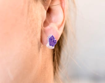 以下が含まれることがあります： 紫色のクリスタルスタッドイヤリングをつけた人の耳のクローズアップ。イヤリングは小さく繊細で、透明なプラスチック製のバックにセットされています。