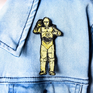 May include: Embroidered patch featuring a yellow and black C-3PO design. The patch is affixed to a light blue denim jacket. The patch is approximately 3 inches tall and 1.5 inches wide.