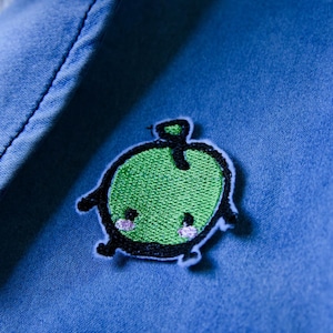 May include: A green apple-shaped patch with a smiling face and black outline is sewn onto blue denim fabric. The patch has a green embroidered body, black legs, and pink cheeks. The apple has a small green stem.