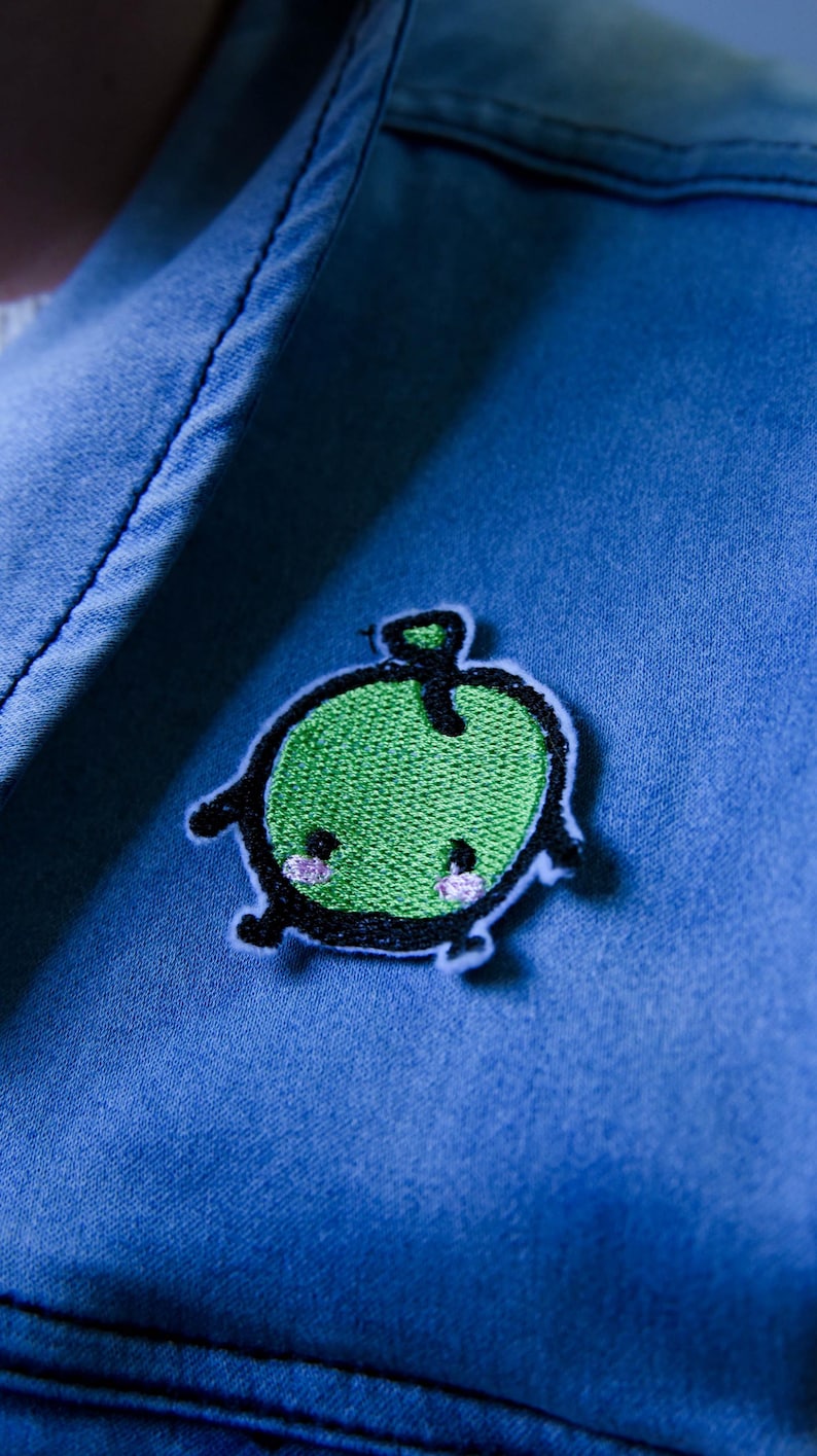 Puede incluir: Un parche verde con forma de manzana con una cara sonriente y un contorno negro est&aacute; cosido sobre tela vaquera azul. El parche tiene un cuerpo bordado verde, patas negras y mejillas rosas. La manzana tiene un peque&ntilde;o tallo verde.