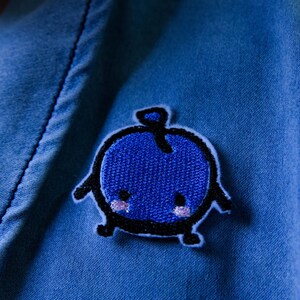 Puede incluir: Un parche bordado azul con forma de ar&aacute;ndano con contorno negro. El ar&aacute;ndano tiene un tallo, brazos y piernas. El parche est&aacute; cosido sobre una tela vaquera azul.