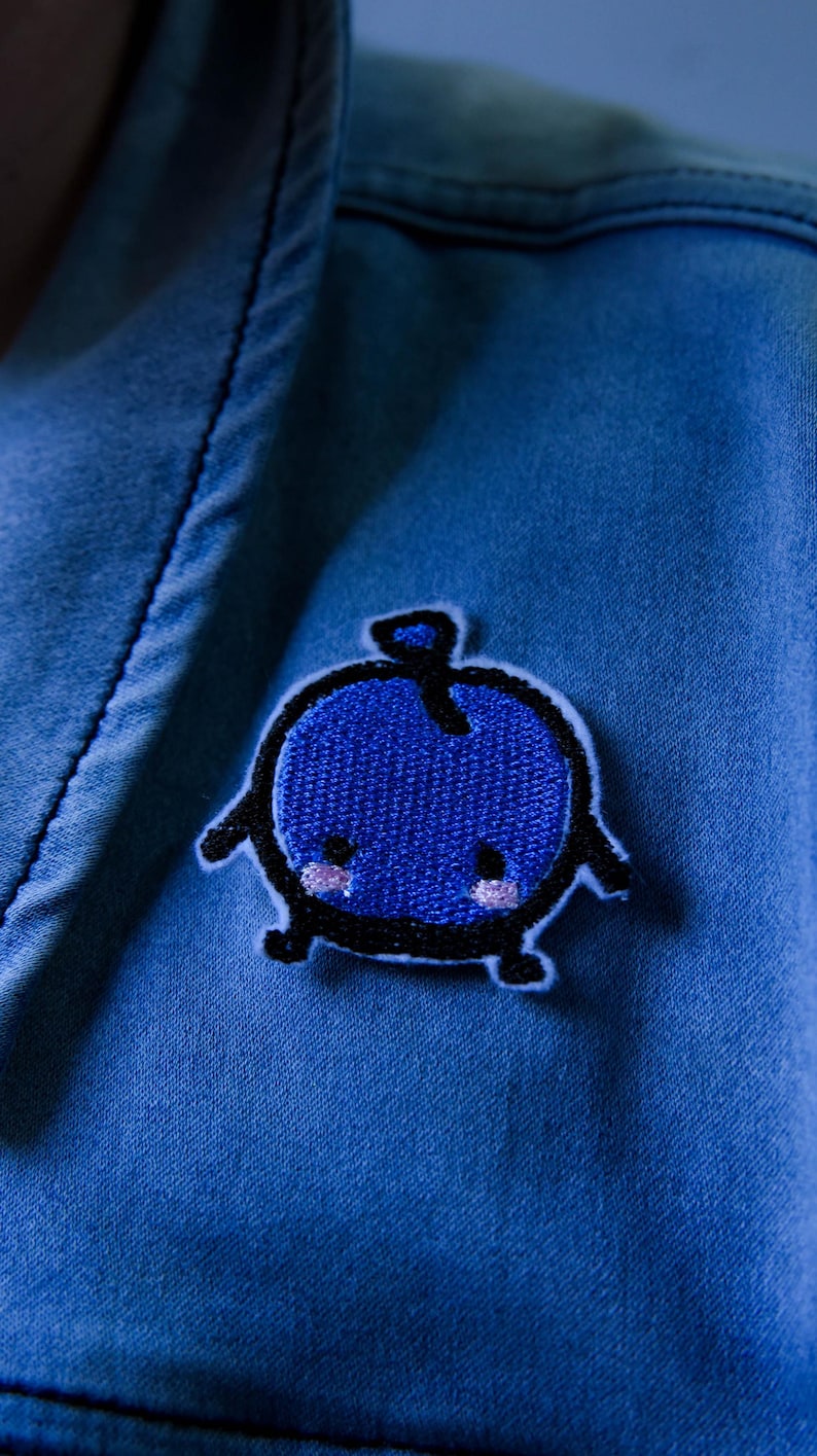 Puede incluir: Un parche bordado azul con forma de ar&aacute;ndano con contorno negro. El ar&aacute;ndano tiene un tallo, brazos y piernas. El parche est&aacute; cosido sobre una tela vaquera azul.