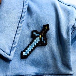 Può includere: Giacca di jeans azzurra con una toppa a spada pixelata nera, blu e gialla. La toppa ricamata è attaccata al petto della giacca. La giacca ha una chiusura con bottoni e un colletto.