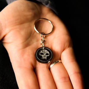 May include: A black keychain with a skull design. The round pendant features a white skull on a black background, attached to a gold-colored keyring. The keychain is held in a hand.