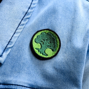 May include: A round, embroidered patch featuring a green tree design on a black background, sewn onto a light blue denim jacket. The tree is detailed with various shades of green, and the jacket has visible stitching.
