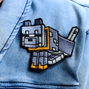 Peut inclure: Écusson brodé représentant un loup pixélisé dans des tons de gris, noir et jaune. L'écusson est cousu sur une veste en jean bleu clair. Le loup est dans une pose ludique, la queue levée.