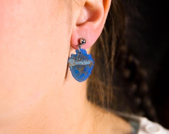 May include: A pair of blue and silver earrings featuring a heart-shaped charm with the word "Ravenclaw" printed on it.