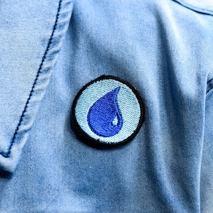 May include: A close-up of a light blue denim jacket with a circular patch. The patch features a blue water droplet design on a light blue background, outlined in black.