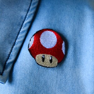 May include: Embroidered red mushroom patch with white spots and a tan base, attached to a light blue denim fabric. The patch is a popular video game character. The denim has visible stitching.