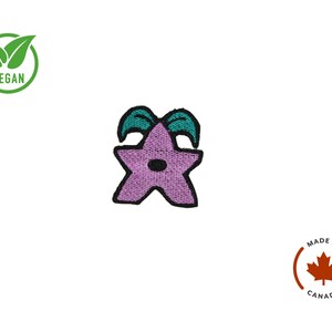 K&ouml;nnte beinhalten: Gestickter Aufn&auml;her mit einem lila Stern mit t&uuml;rkisfarbenen Akzenten und schwarzer Umrandung. Der Aufn&auml;her hat einen kleinen schwarzen Kreis in der Mitte. Das Bild enth&auml;lt auch ein "VEGAN"-Logo und ein "MADE IN CANADA"-Logo.