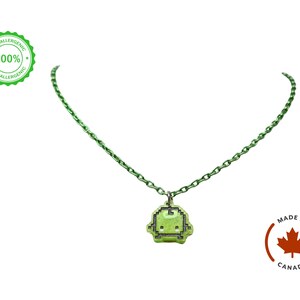 Pu&ograve; includere: Una collana con catena verde con un ciondolo pixelato. Il ciondolo &egrave; di colore verde chiaro con un contorno nero, che ricorda un personaggio di un videogioco retr&ograve;. La collana ha una catena verde abbinata. L'immagine include anche un logo "Made in Canada" e un sigillo "Ipoallergenico".