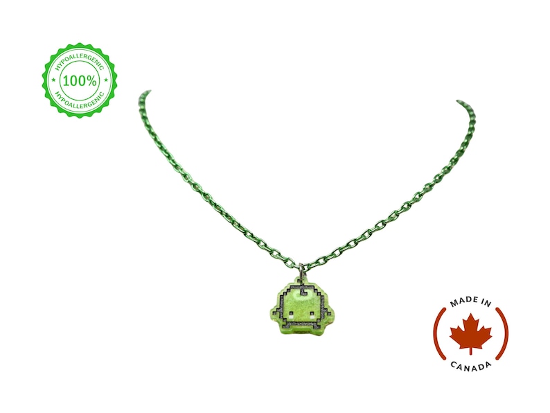 Pu&ograve; includere: Una collana con catena verde con un ciondolo pixelato. Il ciondolo &egrave; di colore verde chiaro con un contorno nero, che ricorda un personaggio di un videogioco retr&ograve;. La collana ha una catena verde abbinata. L'immagine include anche un logo "Made in Canada" e un sigillo "Ipoallergenico".