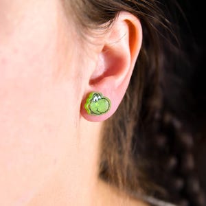 May include: A pair of green and yellow cartoon character stud earrings. The character has a green body and a yellow face.