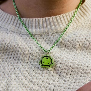 Pu&ograve; includere: Una collana con pendente pixelato verde. Il pendente &egrave; un piccolo design quadrato con un'immagine pixelata di un personaggio. La catena della collana &egrave; anch'essa verde. La collana &egrave; indossata sopra un maglione color crema.