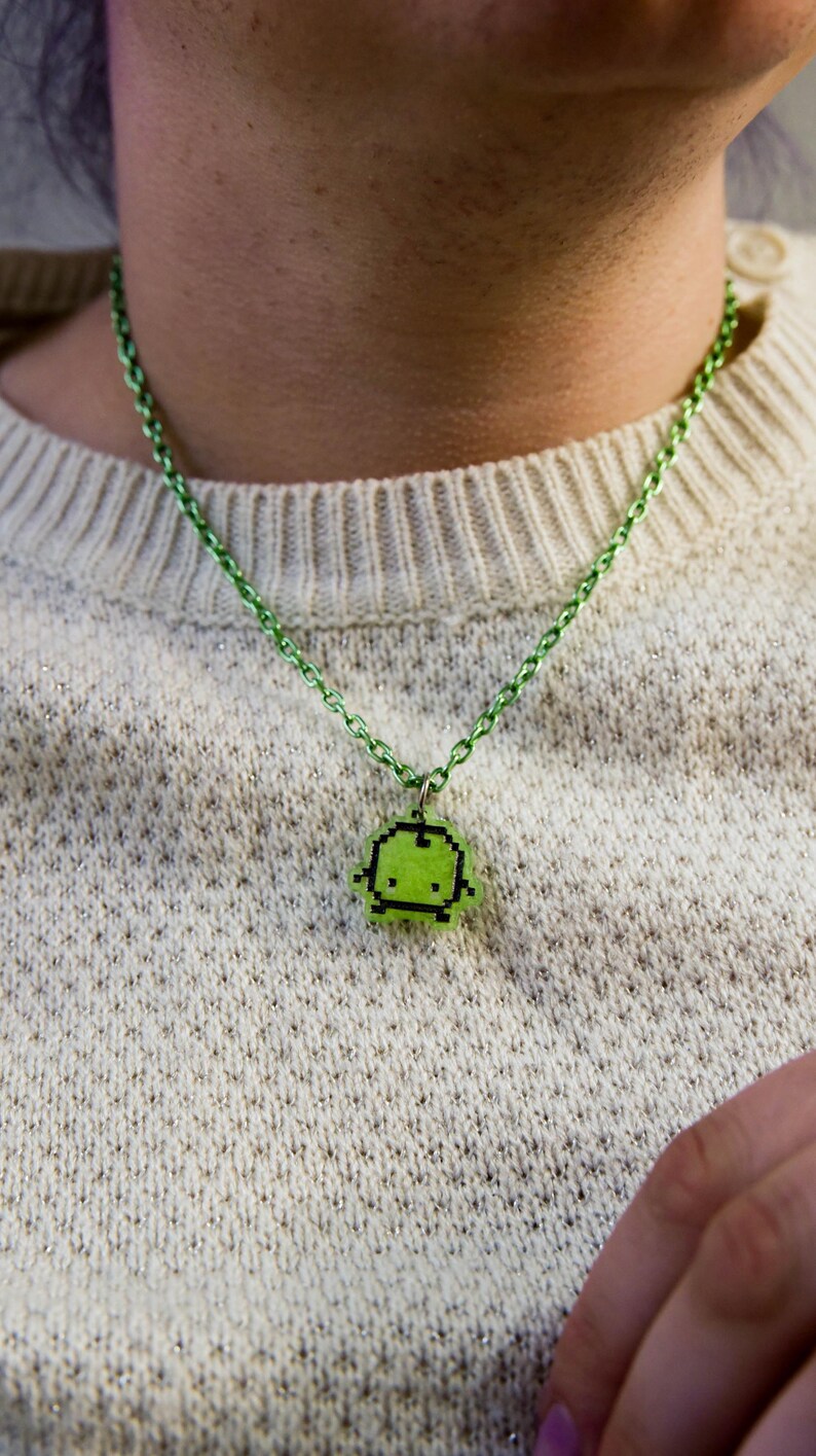 Pu&ograve; includere: Una collana con pendente pixelato verde. Il pendente &egrave; un piccolo design quadrato con un'immagine pixelata di un personaggio. La catena della collana &egrave; anch'essa verde. La collana &egrave; indossata sopra un maglione color crema.