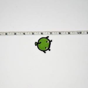 Puede incluir: Un parche verde y redondo con un contorno negro y peque&ntilde;os detalles rosas. El parche mide aproximadamente 2,5 cm de di&aacute;metro. El parche est&aacute; sobre un fondo blanco junto a una regla.