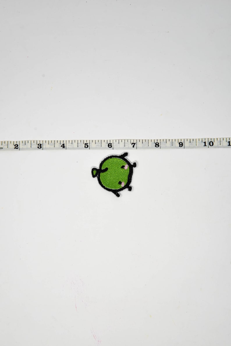 Puede incluir: Un parche verde y redondo con un contorno negro y peque&ntilde;os detalles rosas. El parche mide aproximadamente 2,5 cm de di&aacute;metro. El parche est&aacute; sobre un fondo blanco junto a una regla.