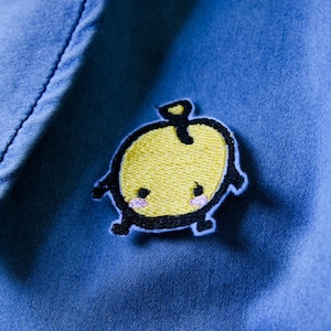 May include: A yellow embroidered apple patch with a black outline and small black legs and arms is sewn onto blue denim fabric. The apple has a small stem and two pink cheeks. The patch is approximately 2 inches wide.