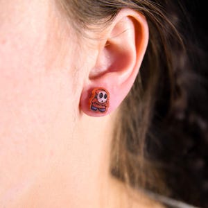 Puede incluir: Primer plano de la oreja de una persona con un pequeño pendiente de un personaje naranja y negro. El personaje lleva una capa roja con capucha y tiene la cara blanca.
