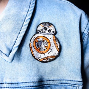 May include: Embroidered BB-8 droid patch, in white, orange, and black, attached to a light blue denim jacket. The patch is a detailed representation of the Star Wars character. The jacket is made of denim.
