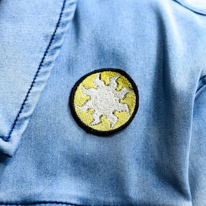 May include: Light blue denim jacket with a round embroidered patch. The patch features a white sun design with yellow rays and a black border. The jacket has a classic collar and dark blue stitching.