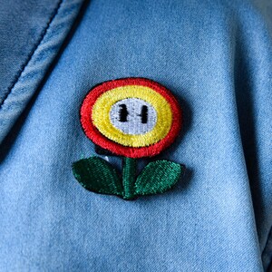 May include: A close-up of a light blue denim jacket with a colorful embroidered flower patch. The patch features a red and yellow flower with a green stem and leaves. The flower has a black and white design in the center.