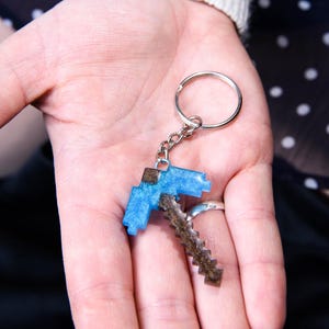 May include: A blue and brown pickaxe keychain with a silver ring. The pickaxe is made of resin and has a glittery finish.