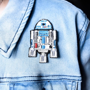 May include: A blue, white, and red embroidered patch of the Star Wars character R2-D2 is affixed to a light blue denim jacket. The patch is rectangular with a detailed design.