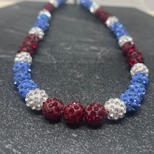 May include: A red, white and blue beaded necklace with a sparkly finish. The beads are arranged in a repeating pattern of red, white and blue.