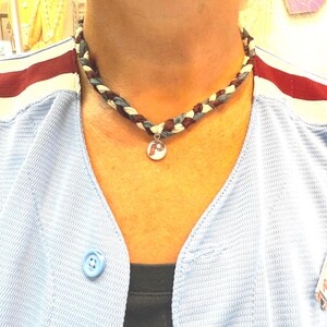 May include: Braided necklace with a pendant featuring a red and white design. The necklace is made of interwoven strands of blue, white, and brown fabric. The pendant has a silver-colored border.