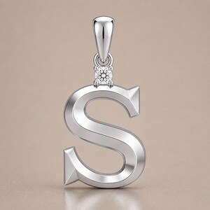May include: A silver pendant in the shape of the letter "S". The pendant has a small, square-cut diamond at the top, where it connects to the bail. The metal has a polished finish, reflecting light. The background is a solid, neutral color.