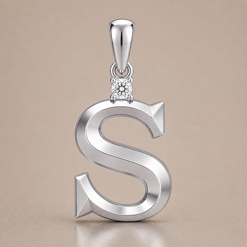 May include: A silver pendant in the shape of the letter "S". The pendant has a small, square-cut diamond at the top, where it connects to the bail. The metal has a polished finish, reflecting light. The background is a solid, neutral color.