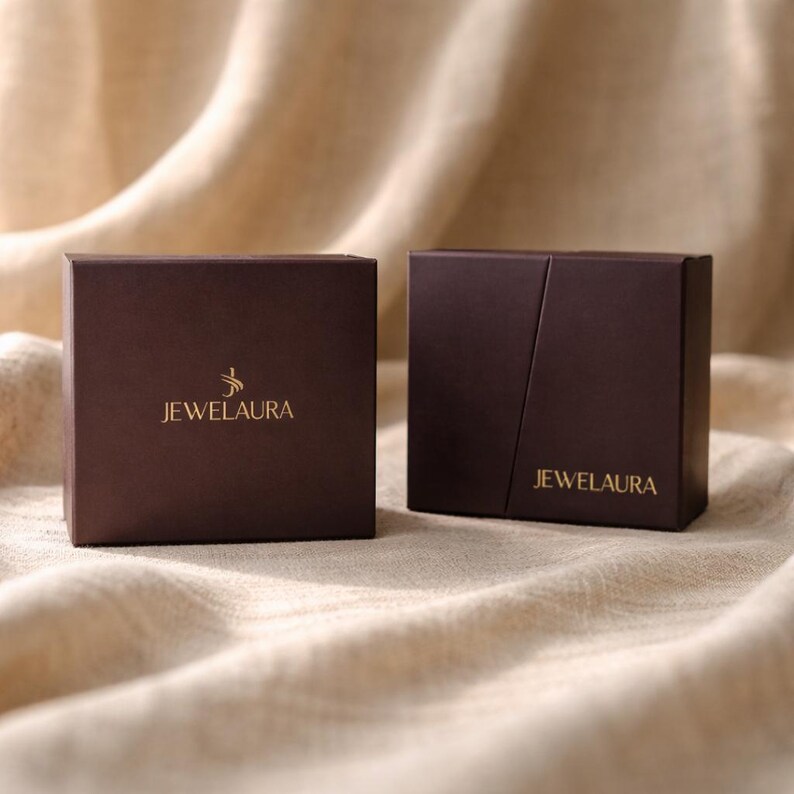 May include: Two brown jewelry boxes with the text "JEWELAURA" in gold lettering. The boxes are square and sit on a beige, textured surface. One box is closed, and the other is open.