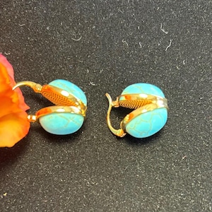 Può includere: Un paio di orecchini dorati con pietre ovali color turchese. Gli orecchini hanno un design a cerniera e sono posti su uno sfondo scuro. Sono un gioiello.