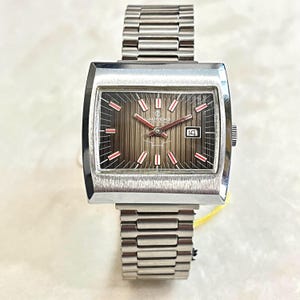 Puede incluir: Un reloj Laurens vintage plateado con una esfera rectangular y una correa plateada. La esfera es marrón con detalles en rojo y blanco, y una pantalla de fecha. La correa es de metal.