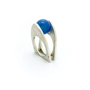 Puede incluir: Un anillo plateado con un diseño moderno, con una esfera azul vibrante sostenida entre dos bandas curvas y abiertas. El anillo tiene una estética minimalista, con un acabado pulido y un contraste llamativo entre el metal y la esfera azul.