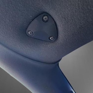 May include: A black triangular cover with three screws, mounted on a blue and black car interior. The cover is located above a round gauge with a black face and white numbers.