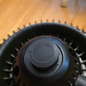 May include: Close-up of a black, circular object with a textured knob on top. The object has a series of small holes and a ridged outer edge. The background is blurred, with a wooden surface visible.
